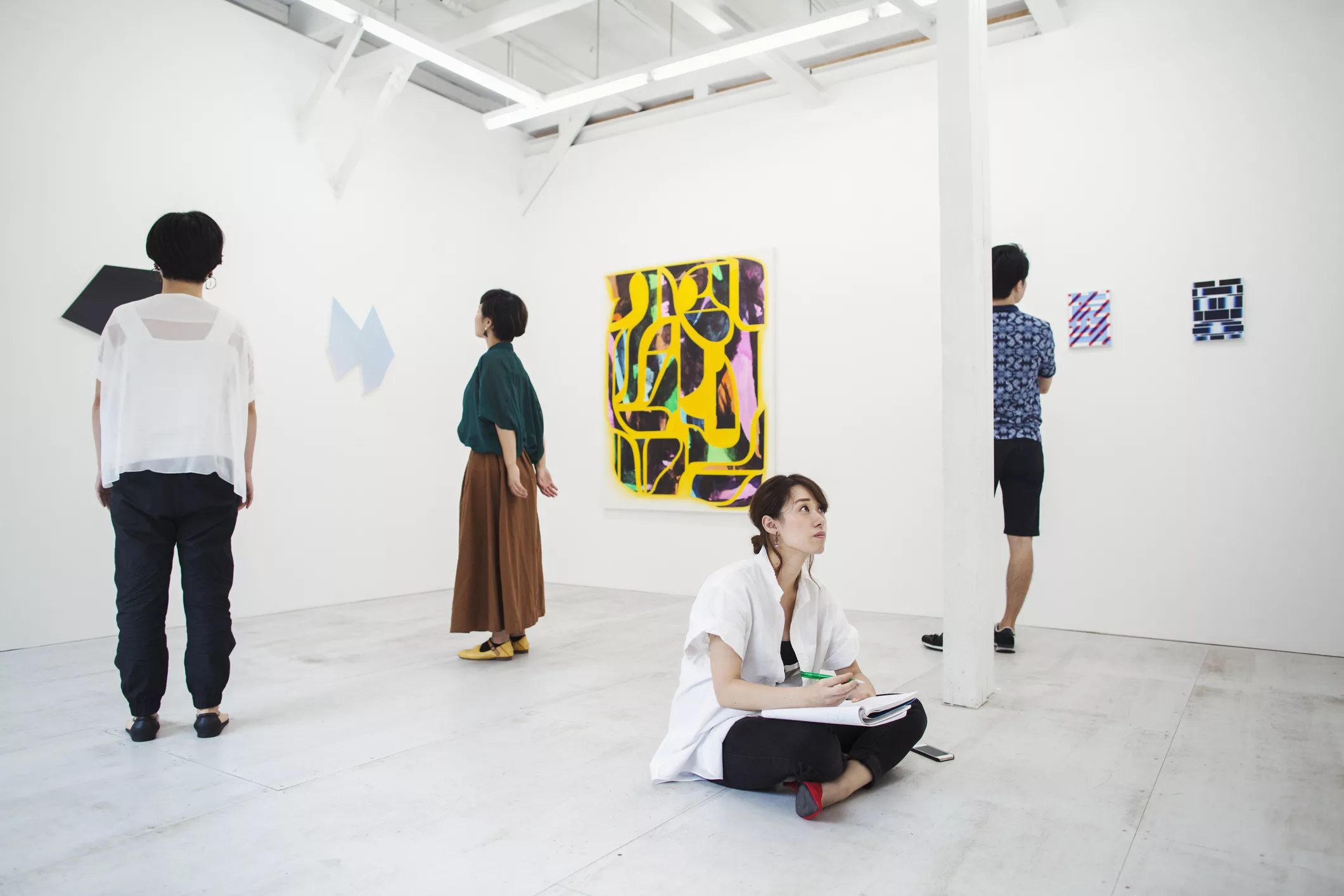 woman-with-black-hair-sitting-on-floor-in-art-gallery-with-pen-and-paper-looking-at-modern-painting-three-people-standing-in-front-of-artworks
