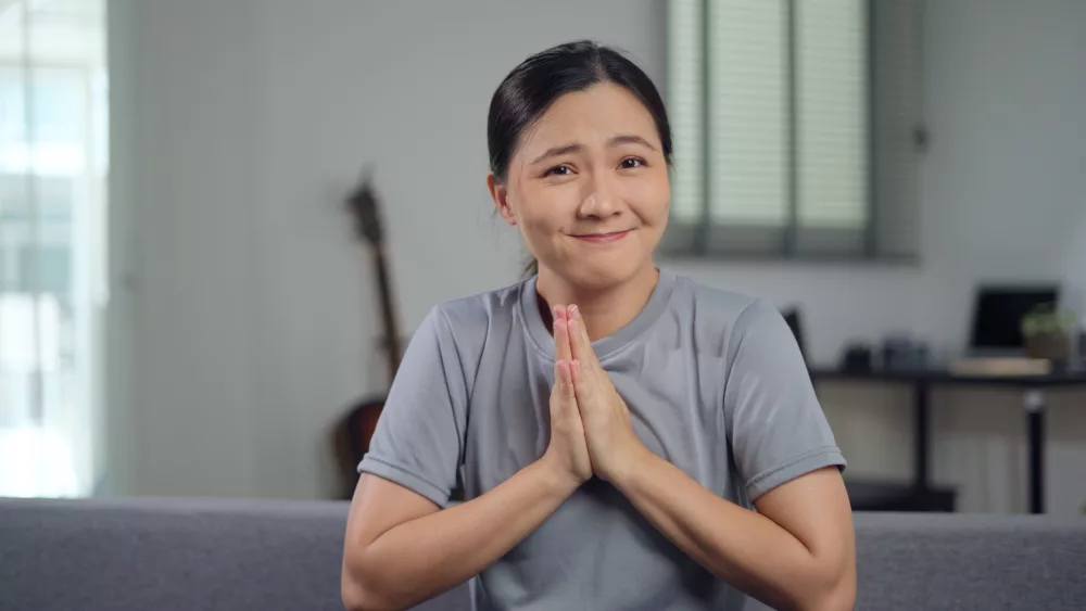 asian-woman-holding-hands-in-prayer-looking-at-camera-sitting-on-sofa-at-home