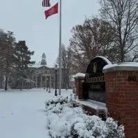 Black-and-white brick entrance sign reading "Monmouth College 1840" in gold letters on a snowy pedestal^ flanked by snow-dusted evergreen bushes. In the background^ a large brick building with a white domed clock tower and arched windows stands amid bare autumn trees under a cloudy gray sky. A tall flagpole flies the U.S. flag and a red Monmouth College banner. The foreground features a snow-covered lawn with scattered evergreens and lamp posts.