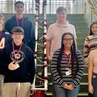Group photo of seven smiling Galesburg High School students selected for the 2026 Illinois All-State music honors^ standing on indoor stairs decorated with red and gold garland and a small lit Christmas tree on the railing. From left to right^ top row to bottom: Mohamed Shehata (top left)^ Carter Bohm (middle left)^ Charlie Ament (bottom left)^ Joshua Cobb (top right)^ Emily Edwards (middle right)^ Jorgiana Donald (bottom middle)^ and Sophia Huizenga (bottom right). Most students are wearing red lanyards with name badges.