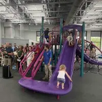 Knox County YMCA CEO Adam Sampson^ in blue^ speaks at a podium as children climb and slide on the new indoor playground during the ribbon-cutting ceremony.