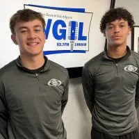 Two Galesburg High School football players in matching gray quarter-zip pullovers stand in front of a WGIL logo screen in the station’s studio