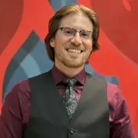 John Glasgow^ smiling^ wearing glasses^ a dark vest^ a burgundy shirt^ and a patterned tie in front of a red and blue mural