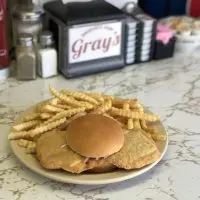 Close-up of a plate on a marble counter holding an oversized breaded pork tenderloin sandwich topped with onions on a toasted bun^ alongside a pile of golden crinkle-cut french fries; in the blurred background are condiments like ketchup^ salt^ and pepper shakers^ plus a napkin holder labeled "Gray's."