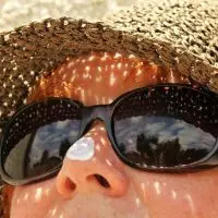 ALT TEXT: Close-up of sunscreen being applied to a person's nose while wearing a sun hat and sunglasses