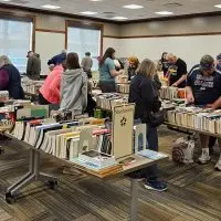 Shoppers browse rows of used books on tables in a large room^ with signs for nonfiction^ local and regional history^ and sports