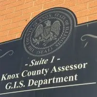 Black sign with the Knox County^ Illinois^ seal reading "Suite 1 – Knox County Assessor^ G.I.S. Department" on a brick wall