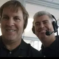Alt text: Jim Lee and Brad Bennewitz smile for a selfie in a broadcast booth^ Bennewitz wearing a headset with a microphone