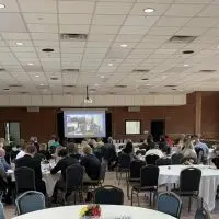 Rows of round tables fill the brick-walled Lake Storey Pavilion as attendees watch a speaker on a large projection screen