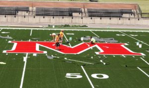 Workers install the new varsity 'M' logo on the Bobby Woll Memorial Field turf on June 12.