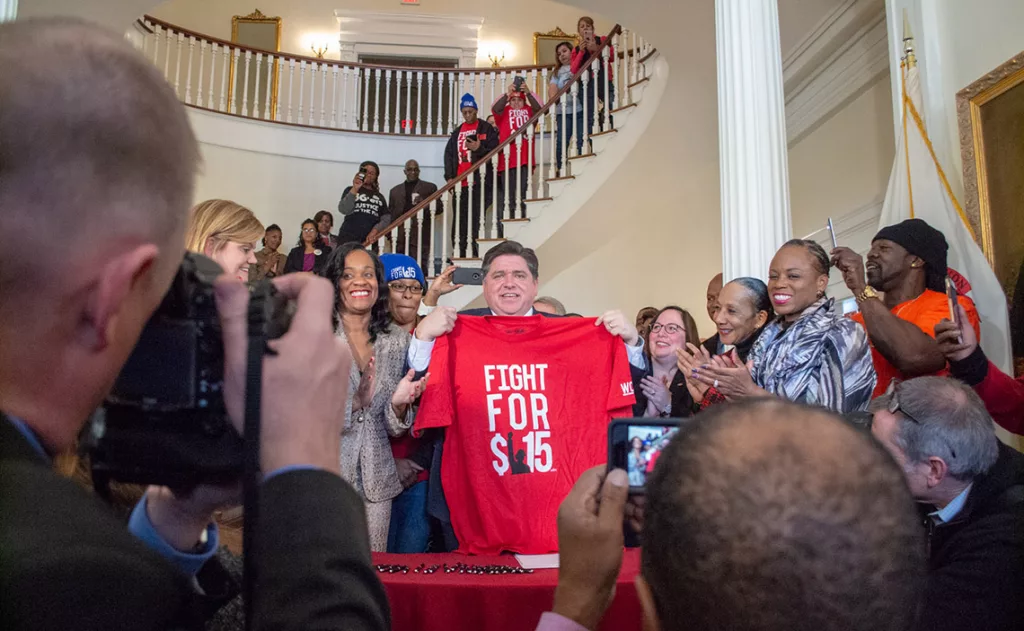 Gov. JB Pritzker holds a shirt commemorating the movement to raise the minimum wage to $15 after signing the bill on February 20, 2019. The wage hits $15 in 2025.