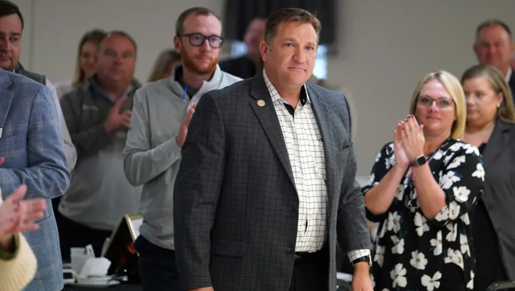 Mark Thomas, wearing a gray plaid suit jacket and white checkered shirt, stands with a humble expression as family, friends, and community members applaud behind him following the surprise announcement of his 2025 Thomas B. Herring Community Service Award at the Galesburg Area Chamber of Commerce Thanksgiving Luncheon.