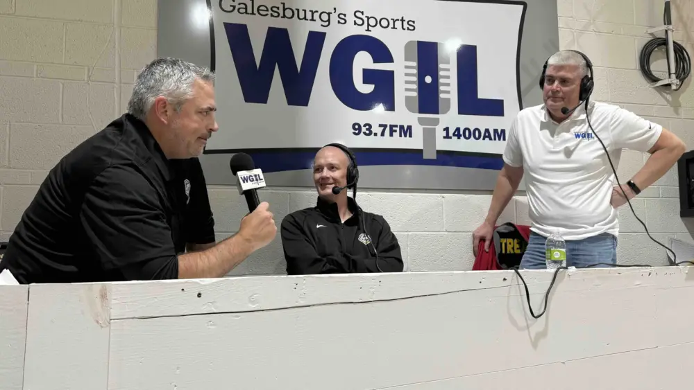 From left: Galesburg head coach Taylor Thiel (seated), color analyst Jason Wessels (seated center with headset), and play-by-play voice Brad Bennewitz (standing right) in front of the WGIL banner at John Thiel Gym.”