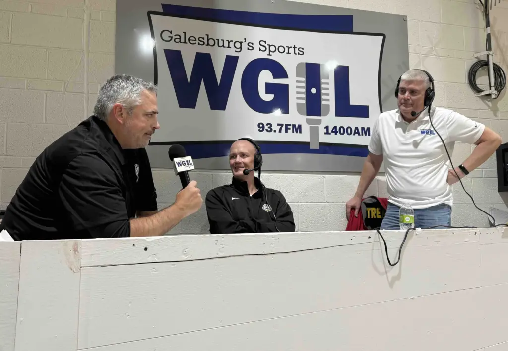 From left: Galesburg head coach Taylor Thiel (seated), color analyst Jason Wessels (seated center with headset), and play-by-play voice Brad Bennewitz (standing right) in front of the WGIL banner at John Thiel Gym.”