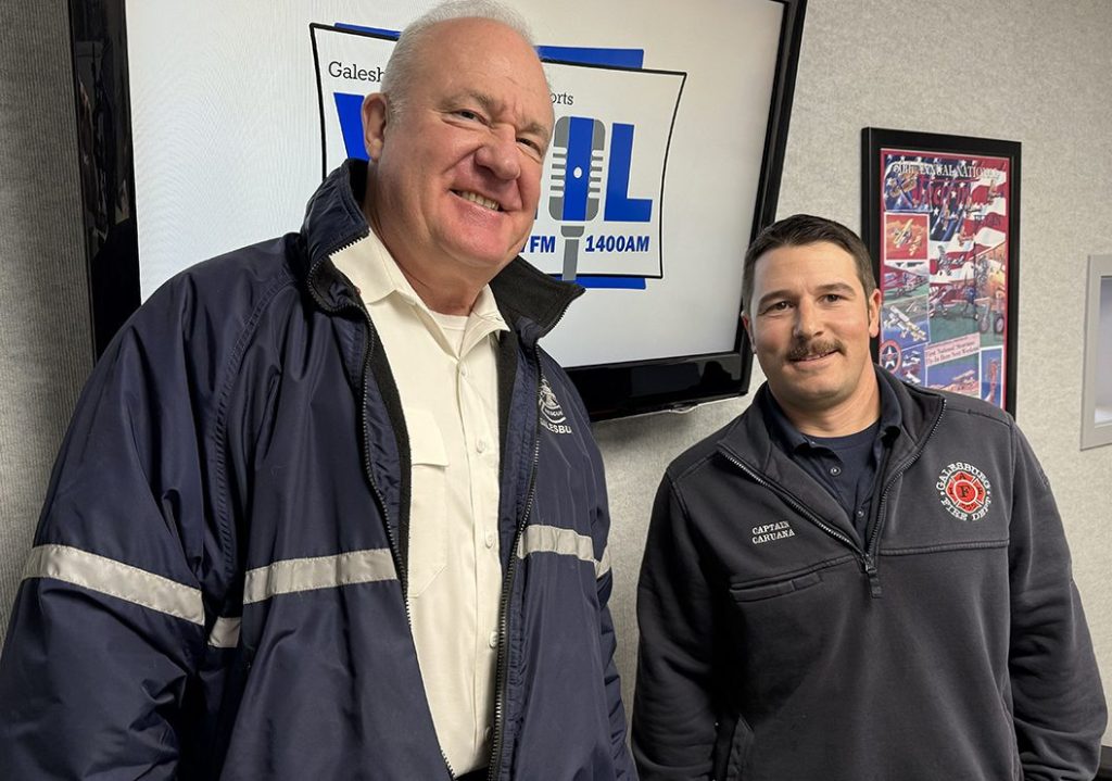 Galesburg Fire Chief Randy Hovind (left, smiling, wearing navy fire department jacket) and Captain Ben Caruana (right, wearing gray Galesburg Fire Department quarter-zip with name tag) pose in the WGIL studio in front of the Galesburg Sports WGIL 1400 AM & 93.7 FM logo.