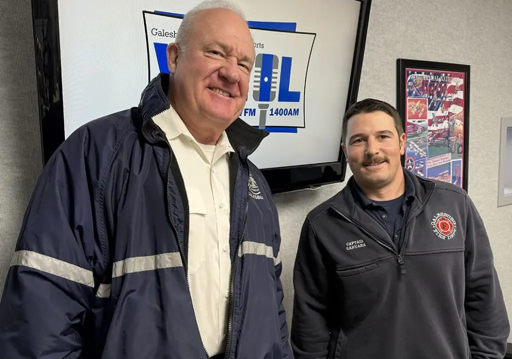 Galesburg Fire Chief Randy Hovind (left, smiling, wearing navy fire department jacket) and Captain Ben Caruana (right, wearing gray Galesburg Fire Department quarter-zip with name tag) pose in the WGIL studio in front of the Galesburg Sports WGIL 1400 AM & 93.7 FM logo.