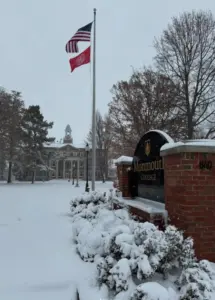 Black-and-white brick entrance sign reading "Monmouth College 1840" in gold letters on a snowy pedestal, flanked by snow-dusted evergreen bushes. In the background, a large brick building with a white domed clock tower and arched windows stands amid bare autumn trees under a cloudy gray sky. A tall flagpole flies the U.S. flag and a red Monmouth College banner. The foreground features a snow-covered lawn with scattered evergreens and lamp posts.