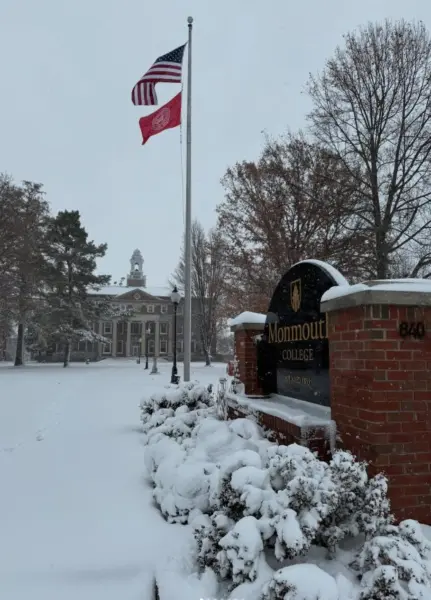 Black-and-white brick entrance sign reading "Monmouth College 1840" in gold letters on a snowy pedestal, flanked by snow-dusted evergreen bushes. In the background, a large brick building with a white domed clock tower and arched windows stands amid bare autumn trees under a cloudy gray sky. A tall flagpole flies the U.S. flag and a red Monmouth College banner. The foreground features a snow-covered lawn with scattered evergreens and lamp posts.