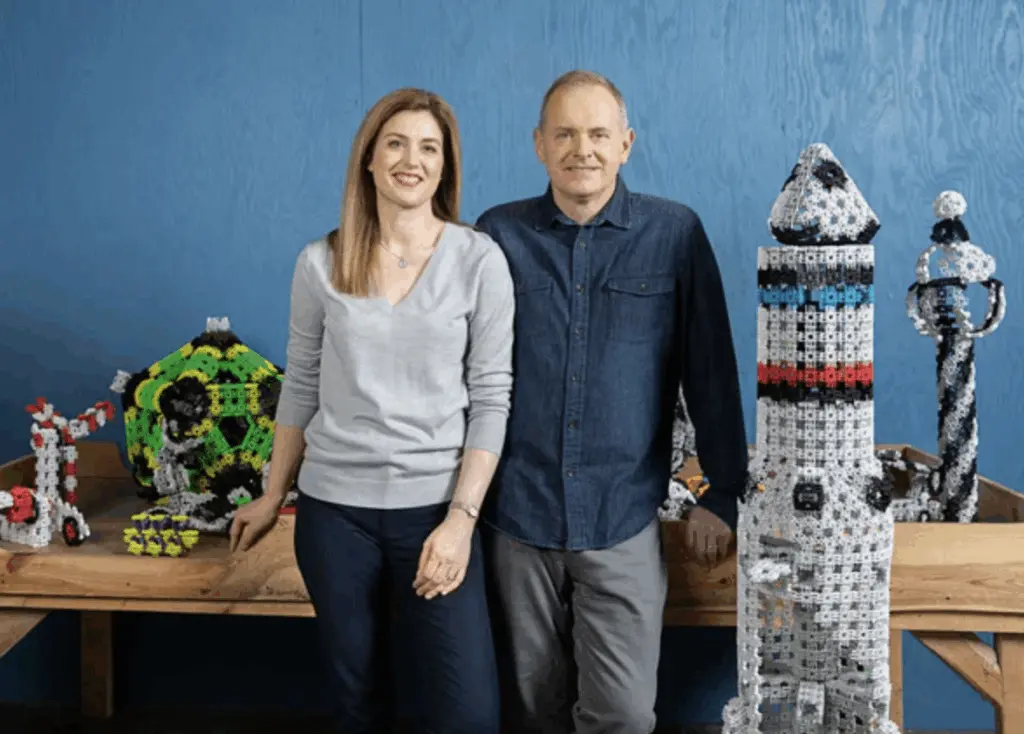 Portrait of Heather Acerra (woman with long blonde hair, smiling, wearing a gray sweater and necklace) and Mike Acerra (man with short hair, smiling, wearing a blue denim shirt), standing arm-in-arm in front of a blue shiplap wall. Surrounding them on a wooden table are colorful Lux Blox magnetic building blocks assembled into a white rocket ship sculpture, green geometric orb, red robot arm, and abstract hinged structures in various shapes and sizes.