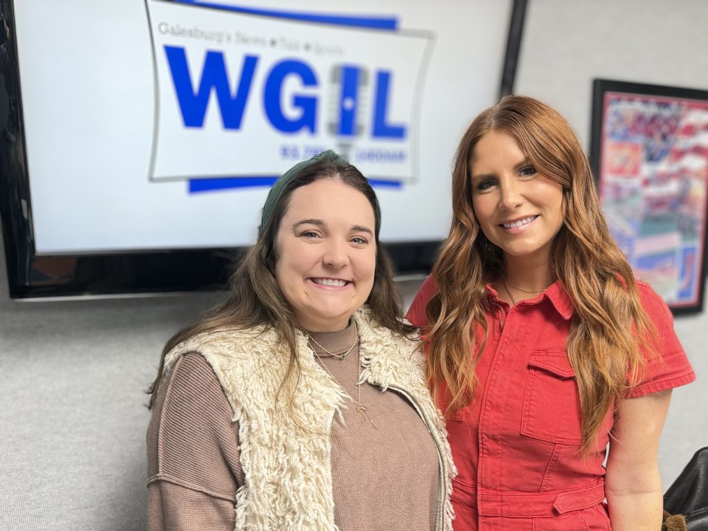 Abby Colvin Thomas (left, wearing green headband and cream shearling vest) and Morgan Seabloom (right, in red jumpsuit) smile in the WGIL studio in front of the WGIL morning news logo.