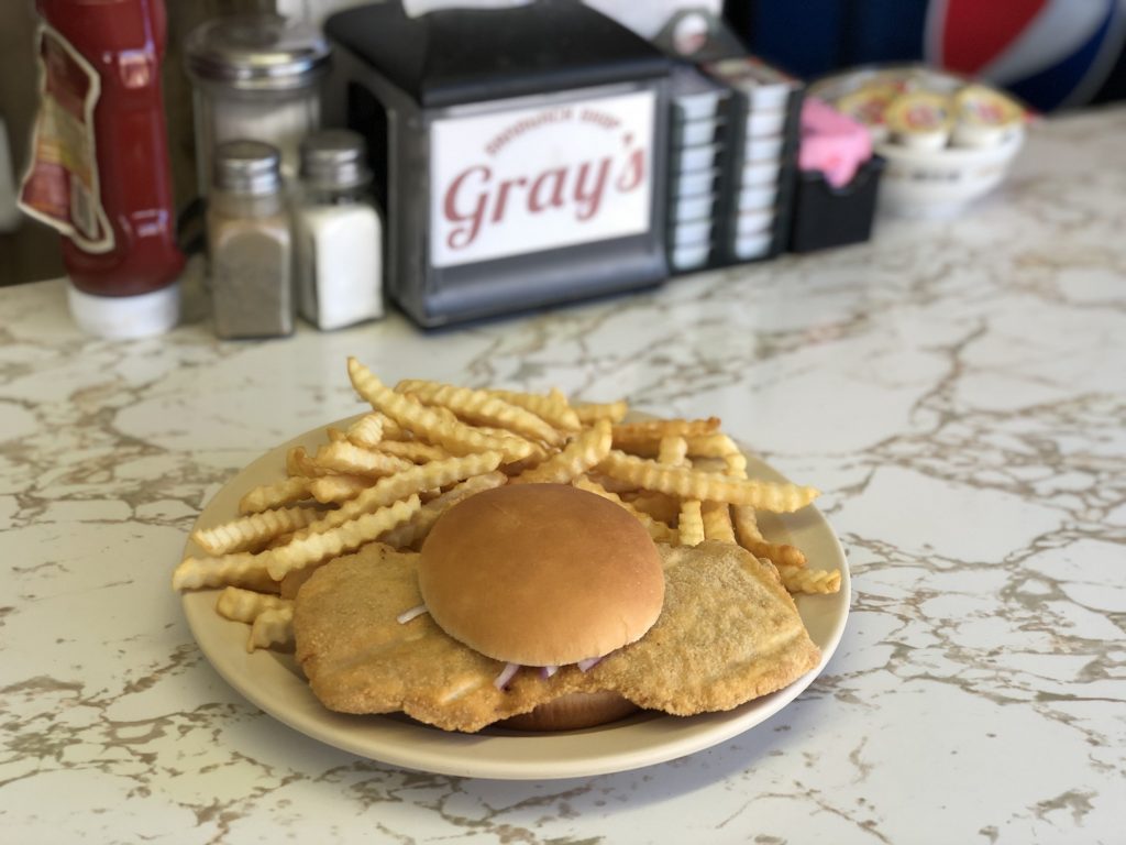 Close-up of a plate on a marble counter holding an oversized breaded pork tenderloin sandwich topped with onions on a toasted bun, alongside a pile of golden crinkle-cut french fries; in the blurred background are condiments like ketchup, salt, and pepper shakers, plus a napkin holder labeled "Gray's."