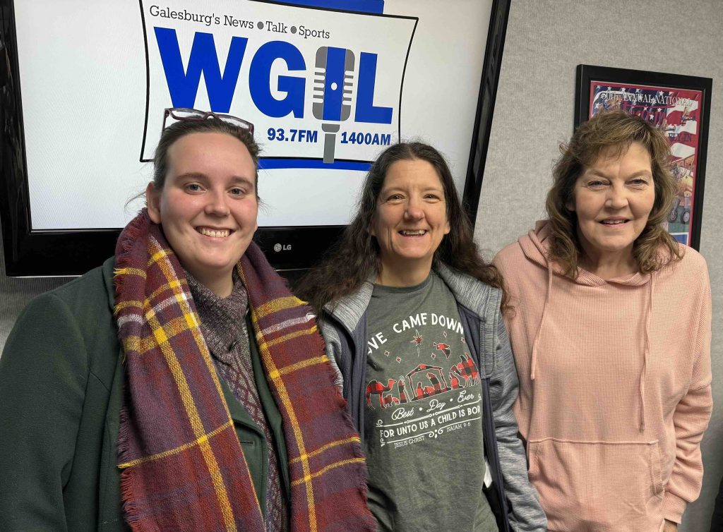 Three smiling women – Choral Dynamics producers Allison Little (left), Deanna Reynolds (center), and Becky Brown (right) – pose together in the WGIL studio in front of the WGIL 93.7 FM • 1400 AM logo, previewing their upcoming holiday show The Beauty of Christmas.