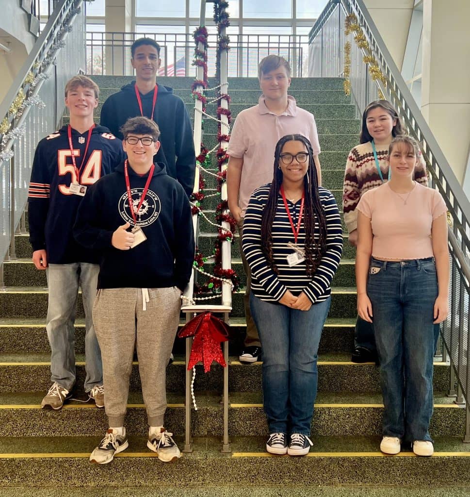 Group photo of seven smiling Galesburg High School students selected for the 2026 Illinois All-State music honors, standing on indoor stairs decorated with red and gold garland and a small lit Christmas tree on the railing. From left to right, top row to bottom: Mohamed Shehata (top left), Carter Bohm (middle left), Charlie Ament (bottom left), Joshua Cobb (top right), Emily Edwards (middle right), Jorgiana Donald (bottom middle), and Sophia Huizenga (bottom right). Most students are wearing red lanyards with name badges.