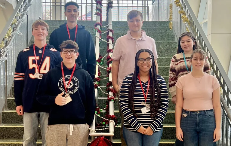 Group photo of seven smiling Galesburg High School students selected for the 2026 Illinois All-State music honors, standing on indoor stairs decorated with red and gold garland and a small lit Christmas tree on the railing. From left to right, top row to bottom: Mohamed Shehata (top left), Carter Bohm (middle left), Charlie Ament (bottom left), Joshua Cobb (top right), Emily Edwards (middle right), Jorgiana Donald (bottom middle), and Sophia Huizenga (bottom right). Most students are wearing red lanyards with name badges.