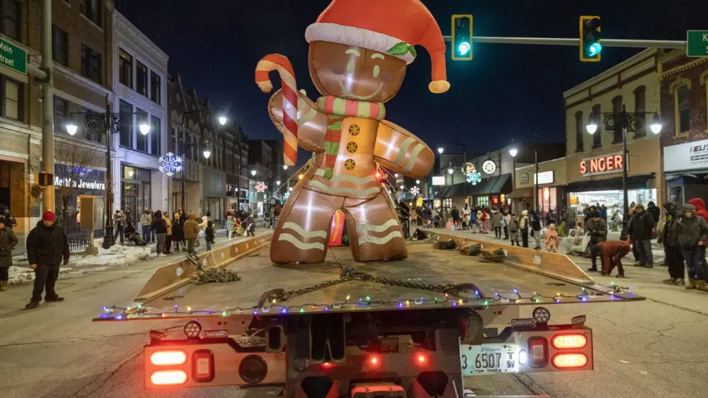 Giant illuminated gingerbread man inflatable on a flatbed trailer rolls through downtown Galesburg during the nighttime Holly Days Parade, with crowds lining both sides of the snow-dusted street and holiday lights glowing in the background. (Photo: Steve Davis/seedcophoto.com)