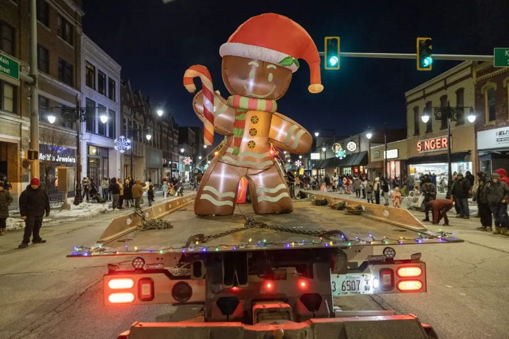 Giant illuminated gingerbread man inflatable on a flatbed trailer rolls through downtown Galesburg during the nighttime Holly Days Parade, with crowds lining both sides of the snow-dusted street and holiday lights glowing in the background. (Photo: Steve Davis/seedcophoto.com)