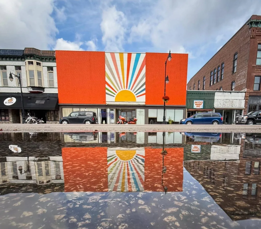 Colorful sunburst mural on the renovated Orange Cup Sidecar building in downtown Galesburg, reflected in a puddle on the street under a partly cloudy sky.