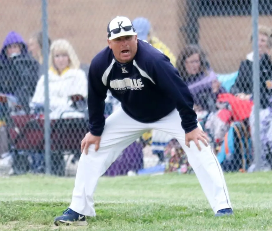 Rick Reed in animated coaching stance on the baseball diamond with spectators behind fence