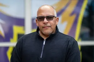 Portrait of Adam Gonzaga, the new head football coach at Knox College, standing in front of the Prairie Fire logo.