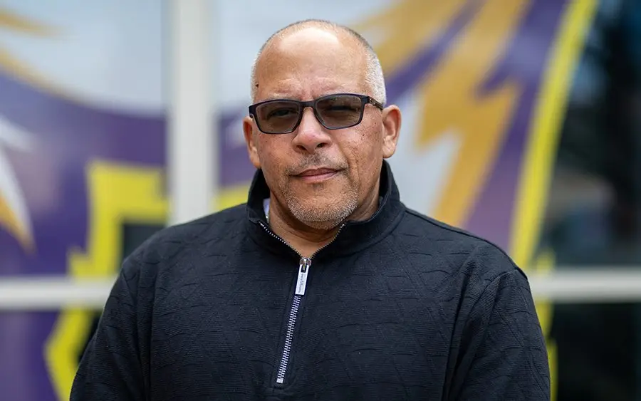 Portrait of Adam Gonzaga, the new head football coach at Knox College, standing in front of the Prairie Fire logo.