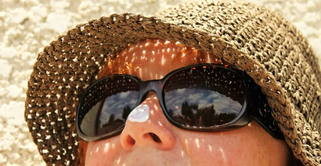 ALT TEXT: Close-up of sunscreen being applied to a person's nose while wearing a sun hat and sunglasses