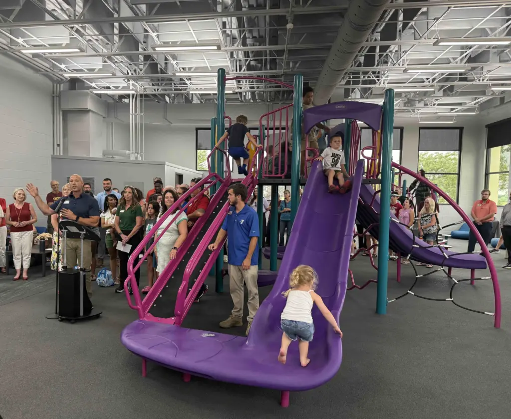 Knox County YMCA CEO Adam Sampson, in blue, speaks at a podium as children climb and slide on the new indoor playground during the ribbon-cutting ceremony.
