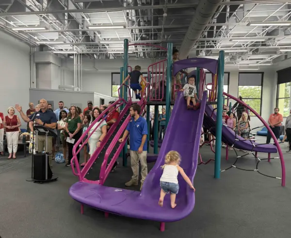Knox County YMCA CEO Adam Sampson, in blue, speaks at a podium as children climb and slide on the new indoor playground during the ribbon-cutting ceremony.