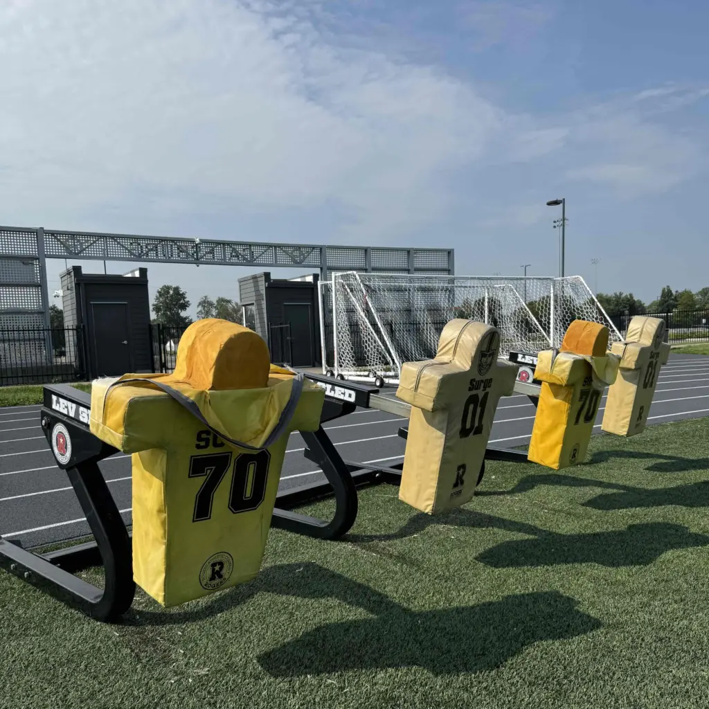 ALT TEXT: Football blocking sleds with padded dummies on the turf at CBI Bank & Trust Stadium