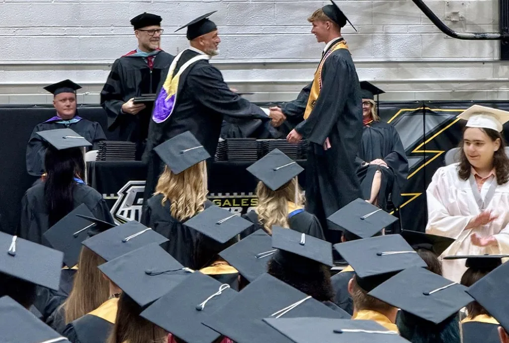 A Galesburg High School graduate in cap and gown shaking hands with a school official while receiving his diploma on stage during commencement