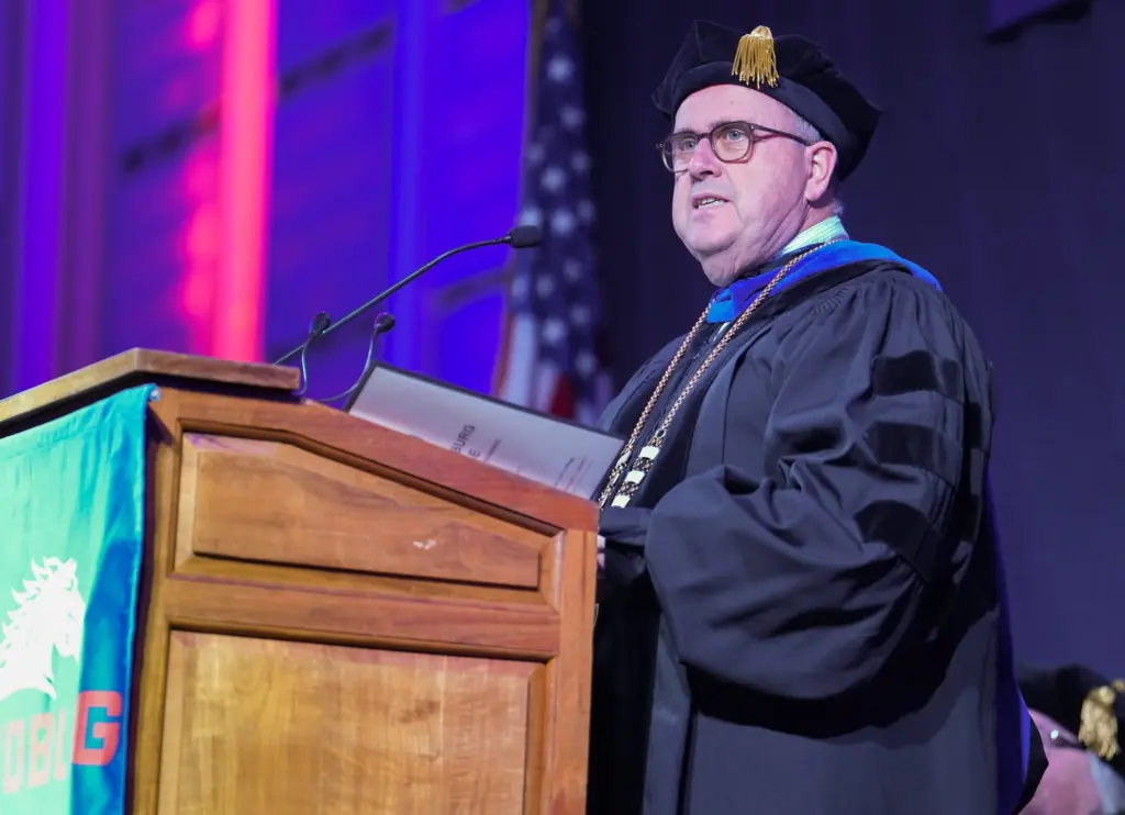 ALT TEXT: Seamus Reilly in academic regalia speaking at a podium during a Carl Sandburg College graduation ceremony