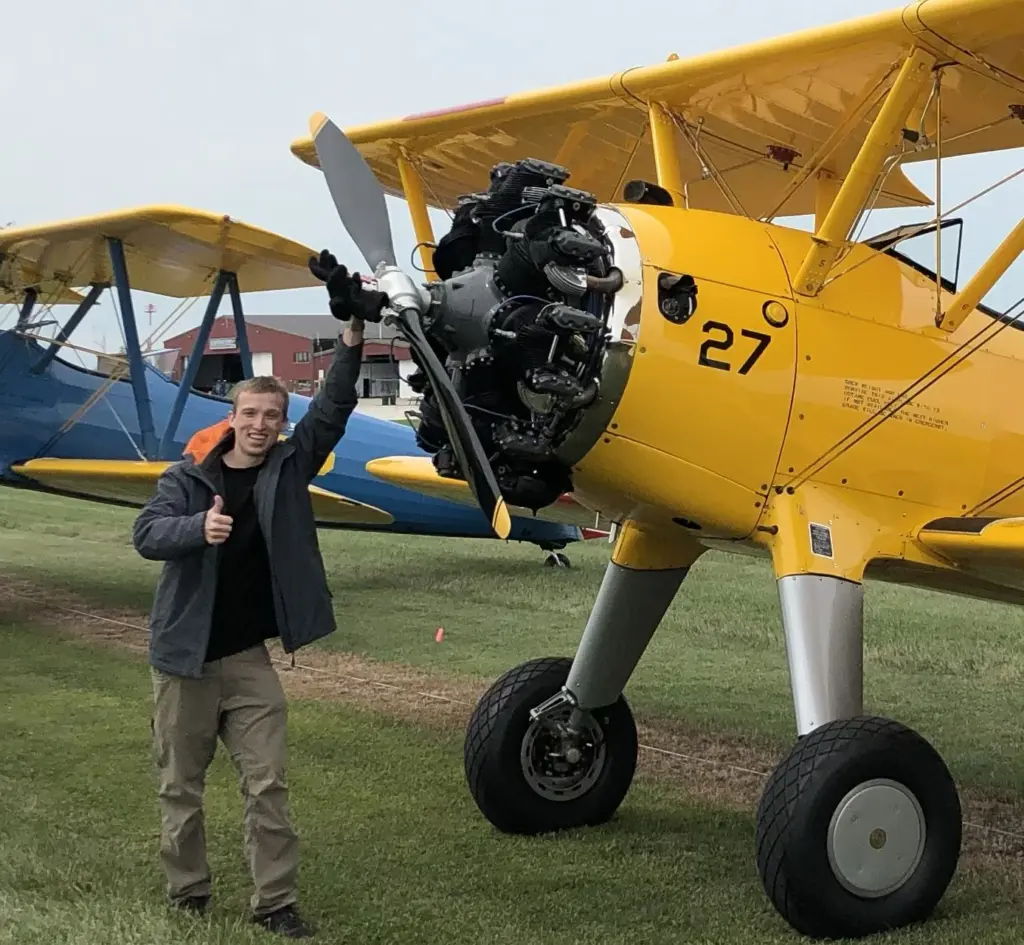 Brett Anderson giving a thumbs up beside a yellow Stearman biplane with its propeller visible