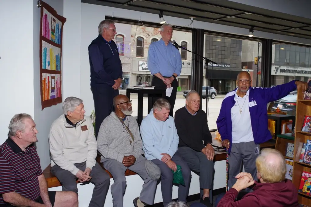 Dale Kelley speaking to a seated crowd at a bookstore event, with two men standing behind him