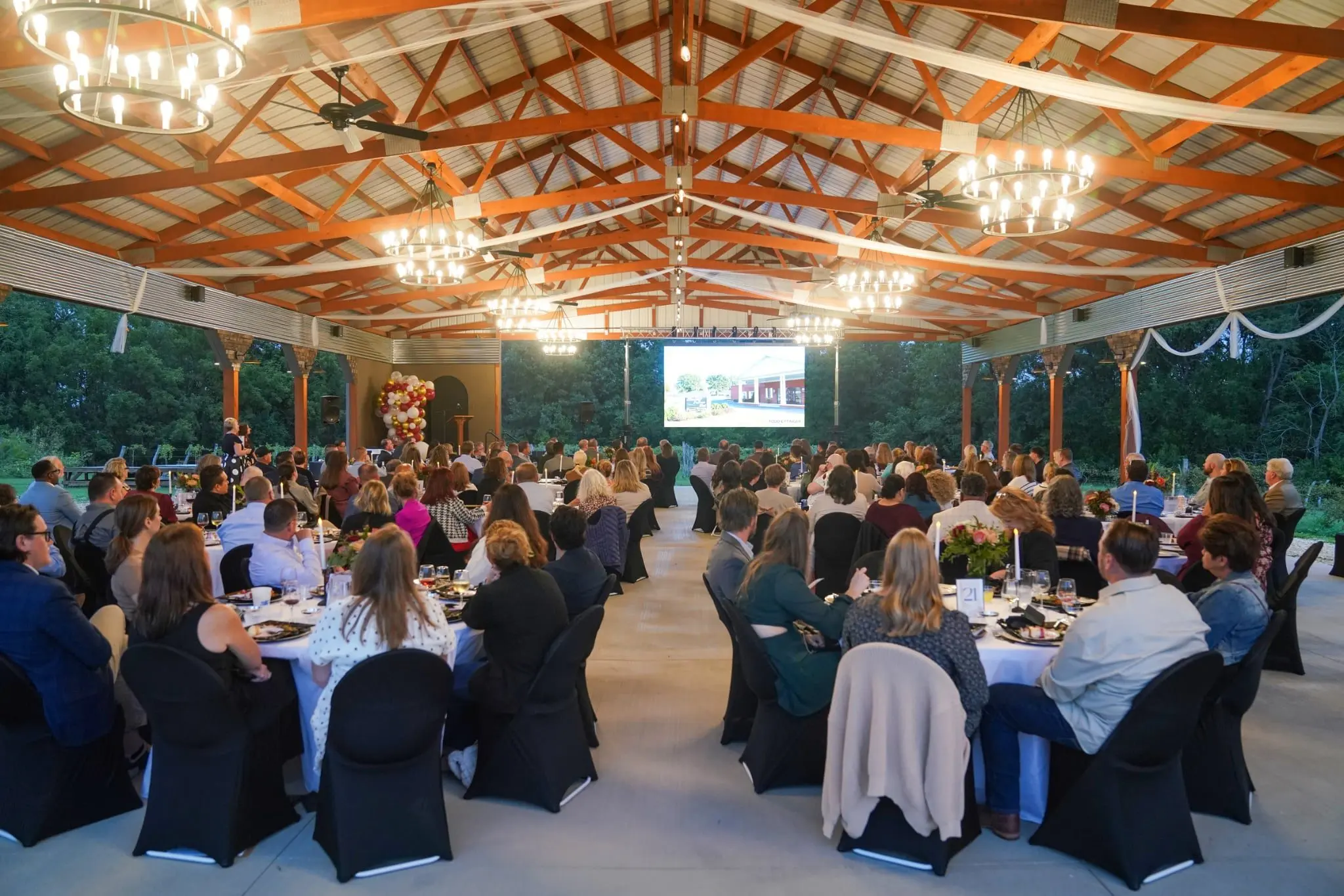 A large crowd seated at tables under a pavilion, watching a presentation screen at a Chamber of Commerce awards event