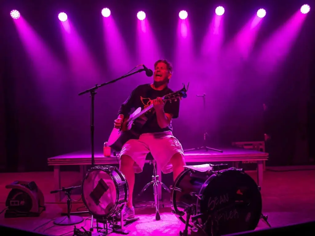 Sean Kistler sings and plays acoustic guitar on a stage lit by purple spotlights, with a kick drum bearing his name in front of him