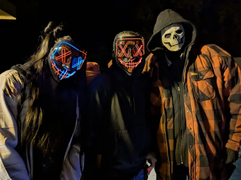 Alt text: Three costumed characters in a dark wooded setting, two wearing glowing LED masks and one in a skull mask and hooded flannel