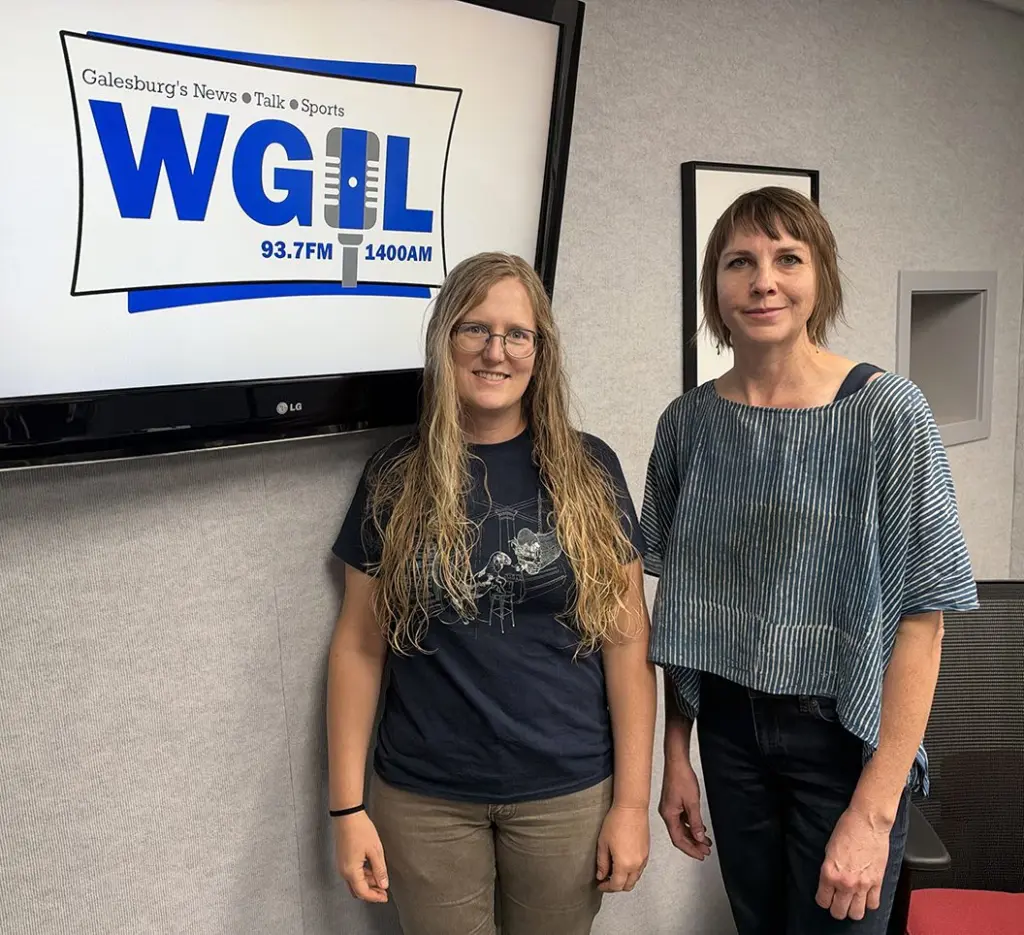 Hannah Lyon and Hillary Anderson stand together beside a screen displaying the WGIL logo