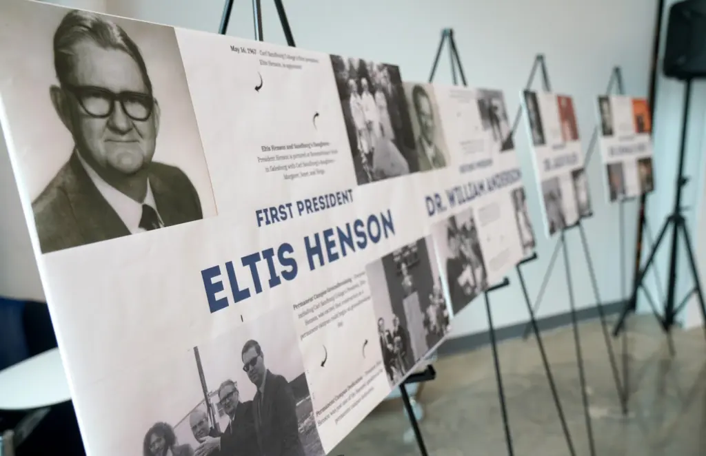 Display board reading "First President Eltis Henson" with a black-and-white portrait of a man in dark-rimmed glasses, beside other presidents' boards on easels