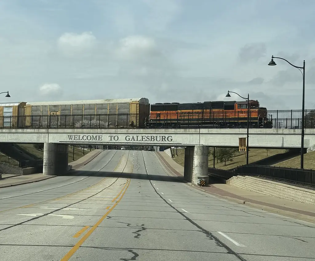 An orange BNSF locomotive pulling enclosed autorack cars across the Sibley Overpass, marked "Welcome to Galesburg," above East Main Street