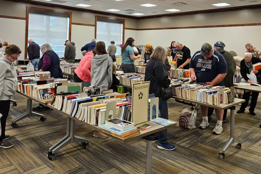 Shoppers browse rows of used books on tables in a large room, with signs for nonfiction, local and regional history, and sports