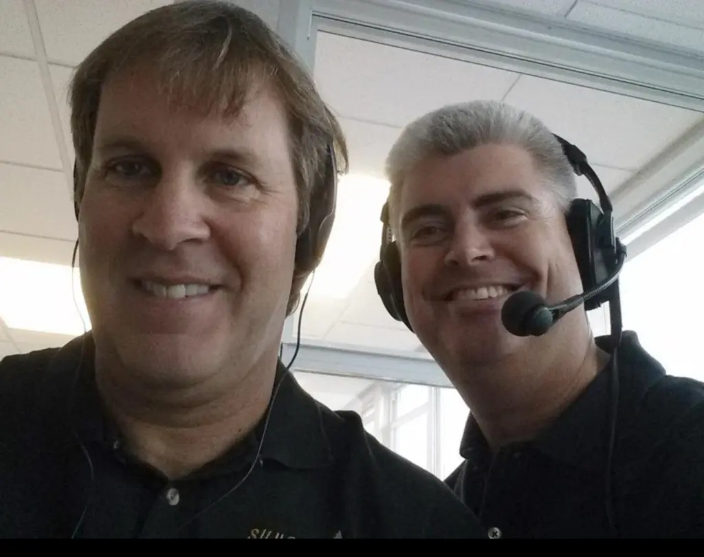 Alt text: Jim Lee and Brad Bennewitz smile for a selfie in a broadcast booth, Bennewitz wearing a headset with a microphone